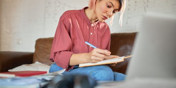 Concentrated young woman copywriter with pink hair working from home making notes in copybook. Cute student girl putting down ideas, writing essay, having focused facial expression, sitting on couch